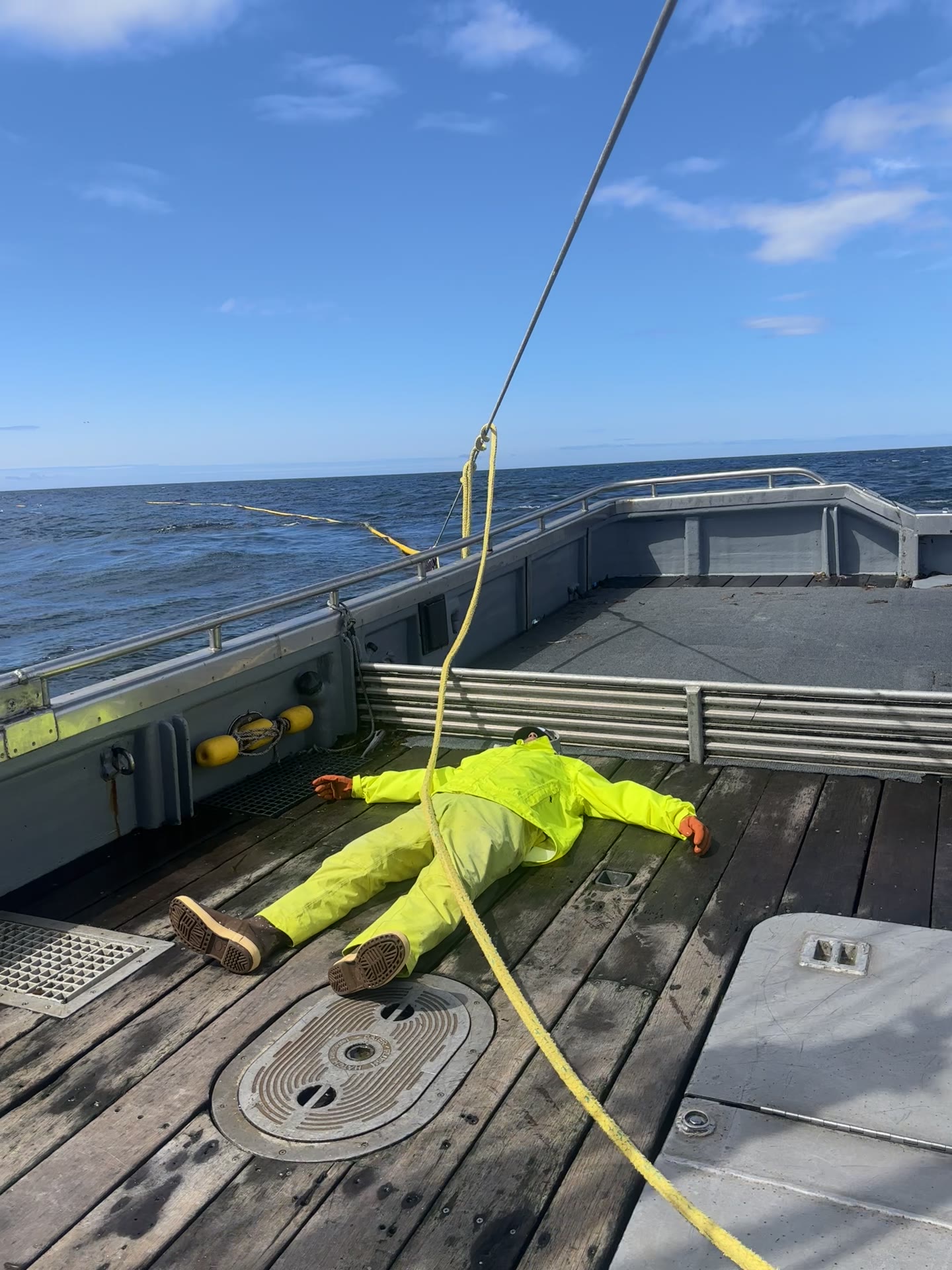 Deckhand in yellow rain gear lying flat on the deck of a seiner under blue sky between sets