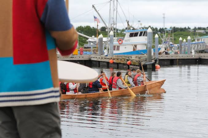 Youth paddling a traditional qayaq out of Sand Point harbor past the fleet